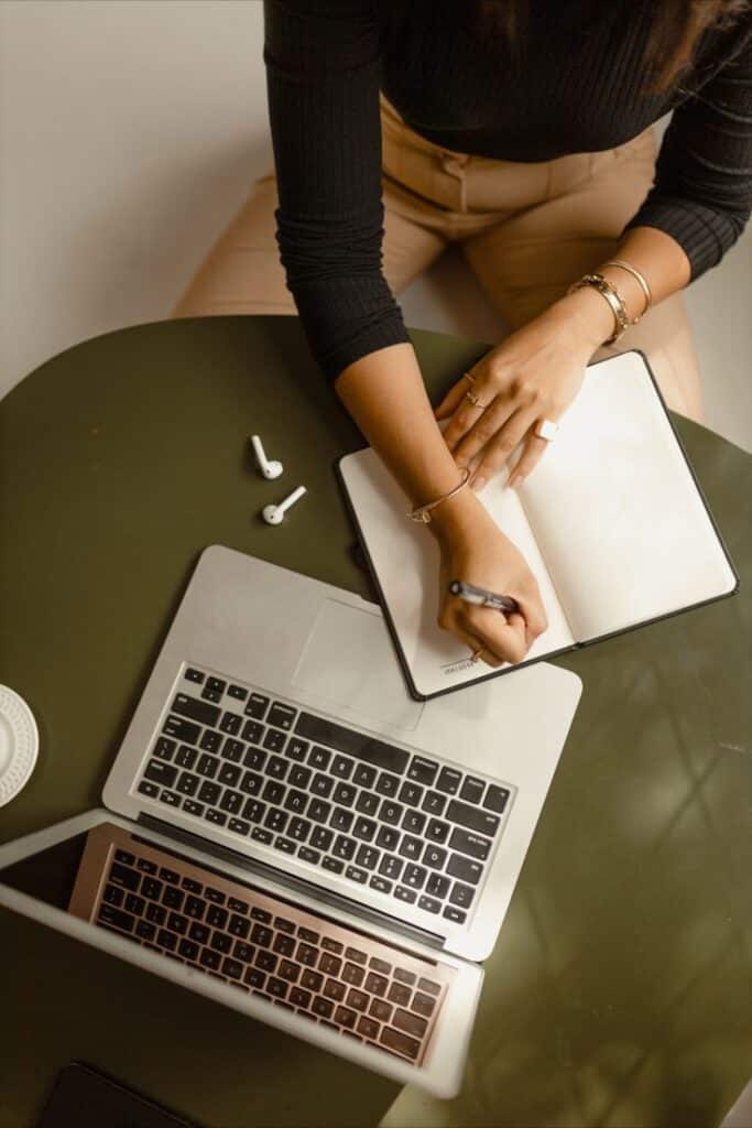 Person writes in notebook beside a laptop on a green desk, wireless earbuds nearby.
