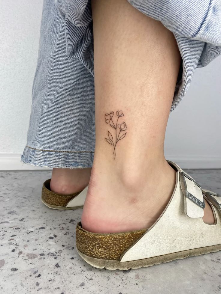 Delicate floral ankle tattoo on person wearing denim jeans and sandals. Perfect minimalist tattoo design.