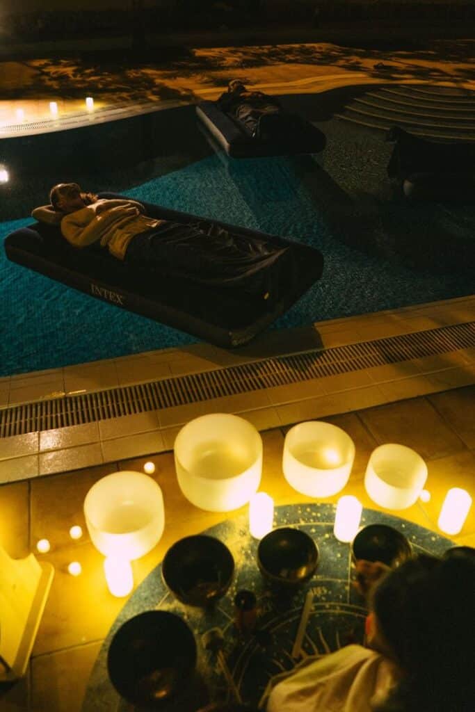 People float on inflatable beds in a pool with lit candles nearby. Relaxing nighttime scene.