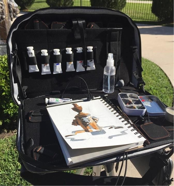 Portable art kit with painting supplies and sketchbook on a sunny day outside, featuring watercolor tubes and brushes.