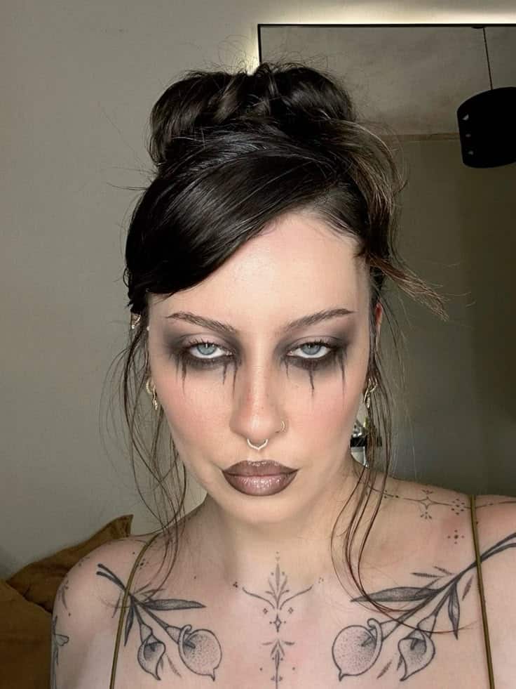 Person with gothic makeup and tattoos poses with a neutral expression; hair is styled in a loose updo.