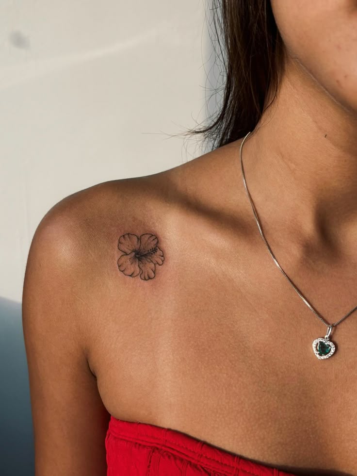 Small flower tattoo on shoulder with heart necklace and red top.