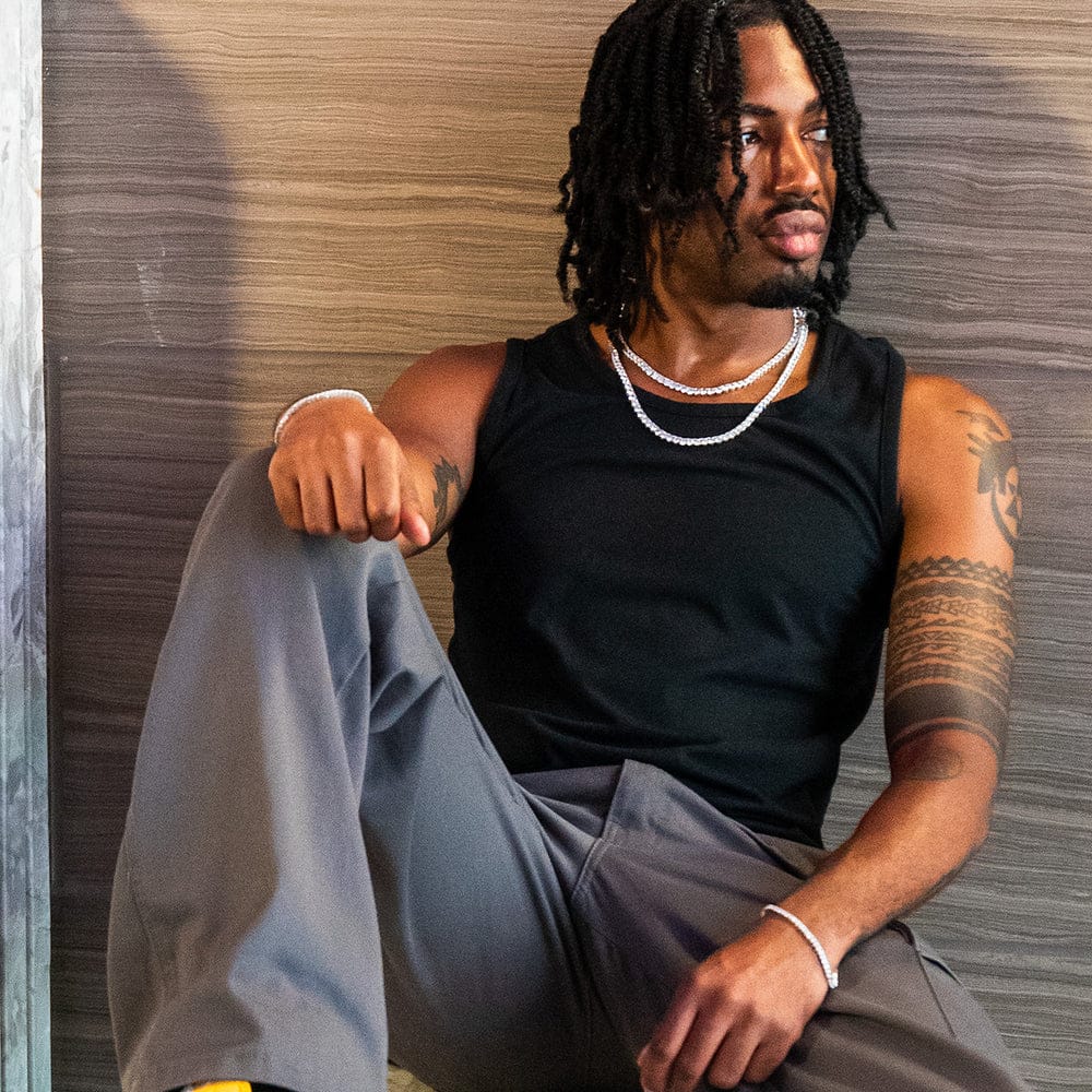 Man with dreadlocks in black tank top and gray pants sitting against a wooden wall, wearing silver jewelry.