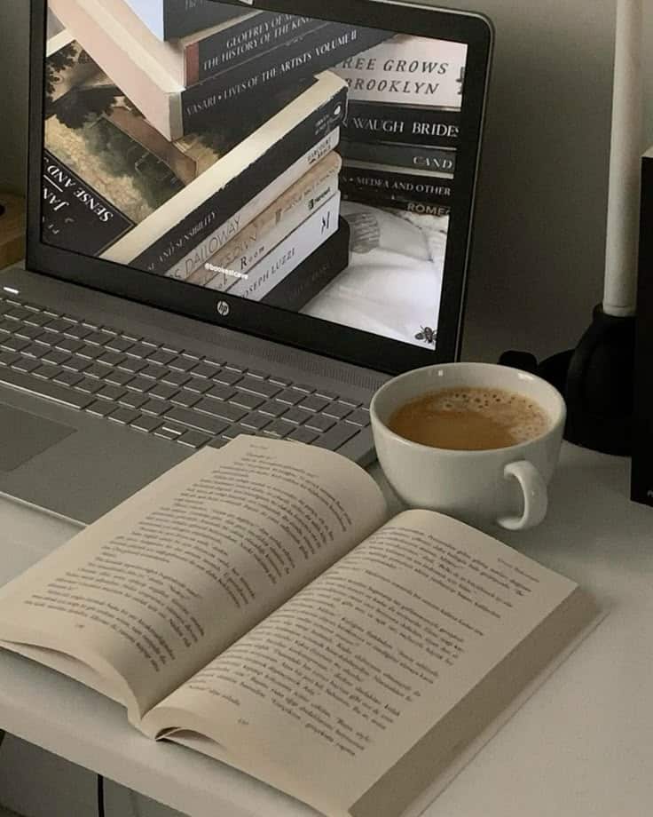 Open book beside a laptop displaying stacked books and a coffee cup, creating a cozy reading atmosphere.
