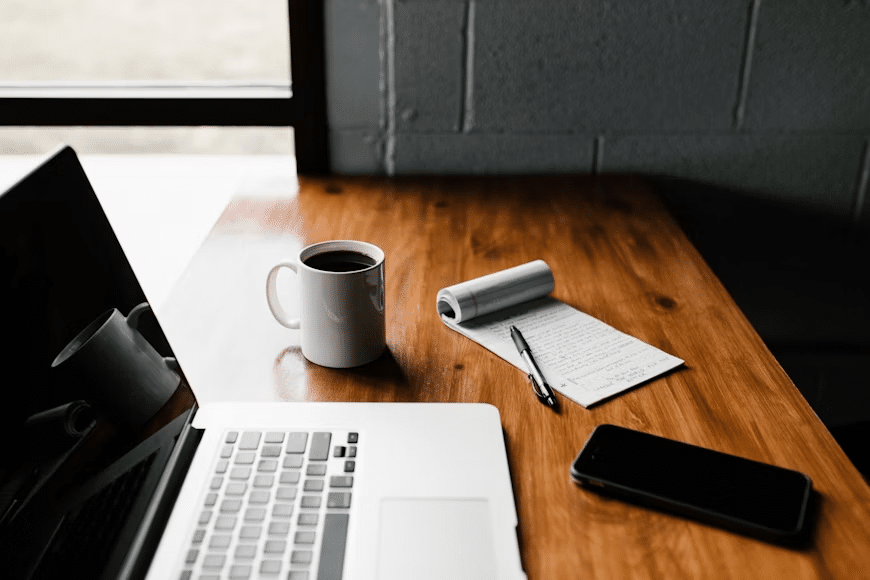 Laptop, coffee mug, notepad, pen, and smartphone on a wooden desk near a window, creating a productive workspace atmosphere.