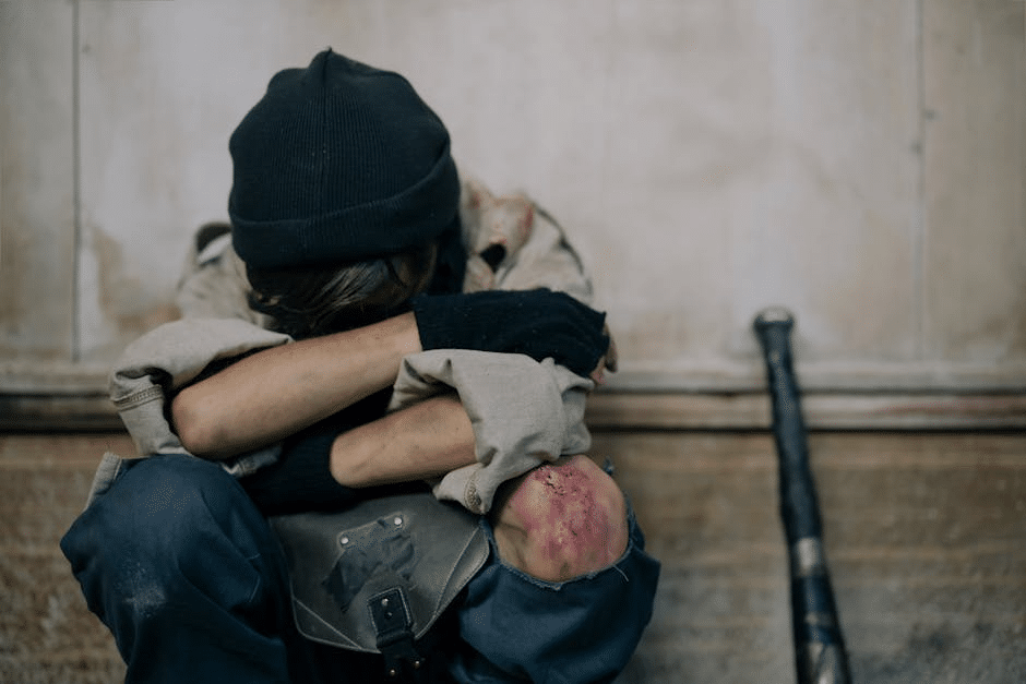 Person sitting against wall, knees drawn up, wearing gloves and beanie, with bat in background.