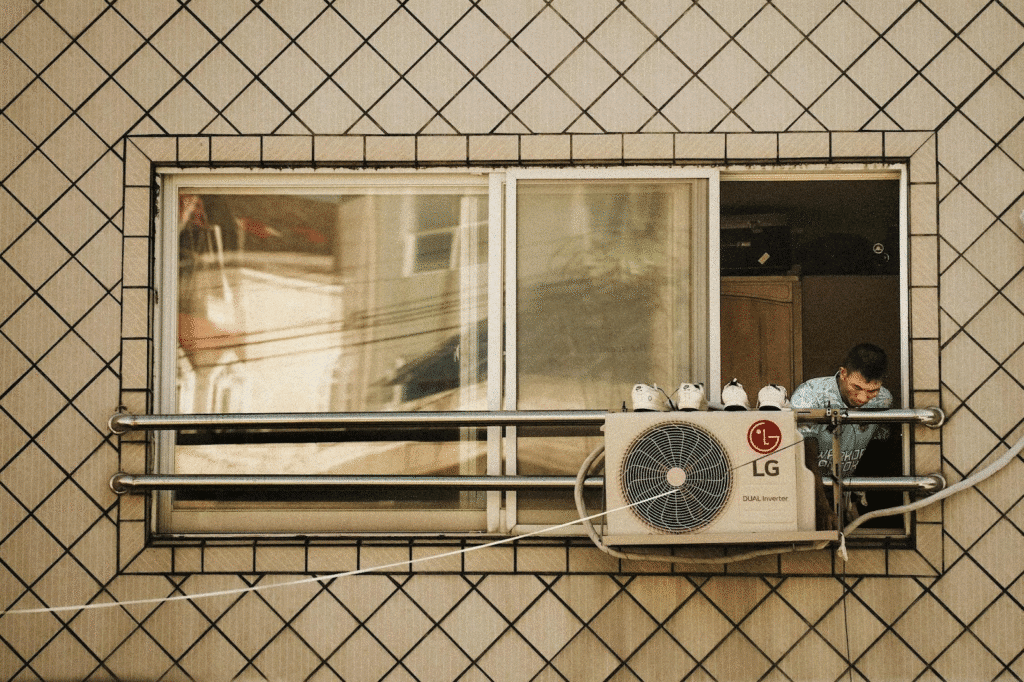 Man leans out window near air conditioner, shoes drying on top, against checkered wall background.