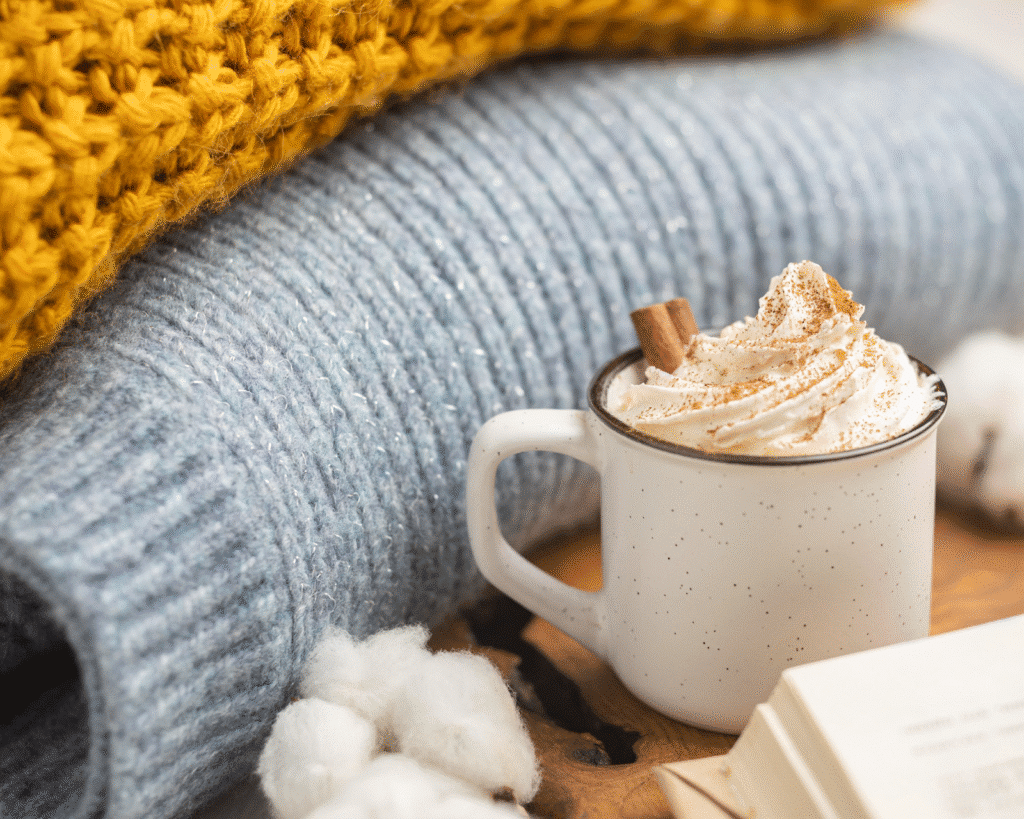 image Cup of whipped cream-topped hot drink with cozy sweaters and book, creating a warm and inviting autumn atmosphere. | Sky Rye Design Cup of whipped cream-topped hot drink with cozy sweaters and book, creating a warm and inviting autumn atmosphere.