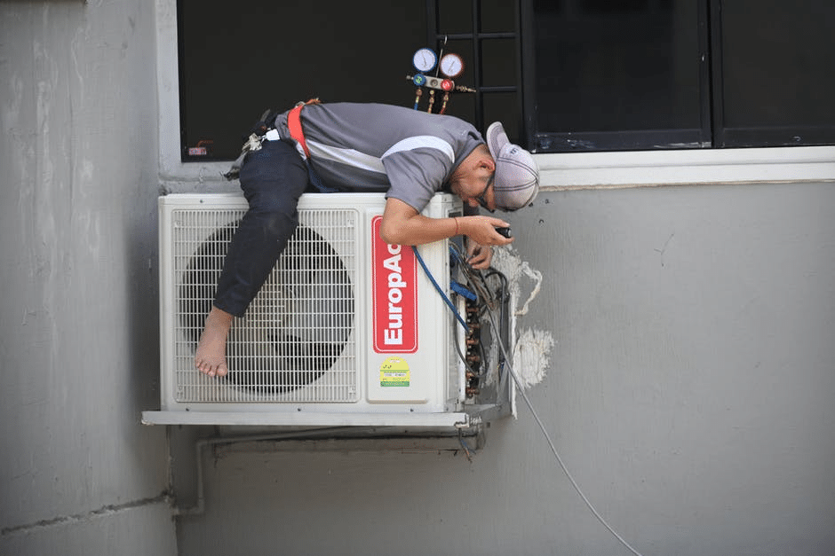 image Technician repairing air conditioner unit on building exterior, using tools and safety harness for maintenance work. | Sky Rye Design Technician repairing air conditioner unit on building exterior, using tools and safety harness for maintenance work.