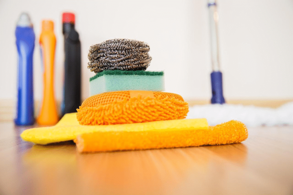 Cleaning supplies stacked on wooden floor, including sponges, scrubbers, and cleaning bottles in the background.