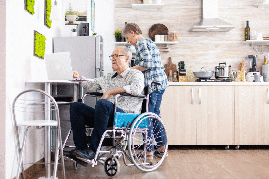 Senior man in wheelchair using laptop while woman cooks in modern kitchen.