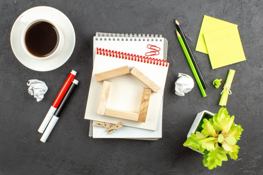 Office desk with coffee, notepads, pens, sticky notes, and a plant arranged creatively on a dark surface.