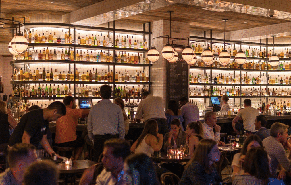 Vibrant bar scene with patrons enjoying drinks, surrounded by illuminated shelves of various bottles.