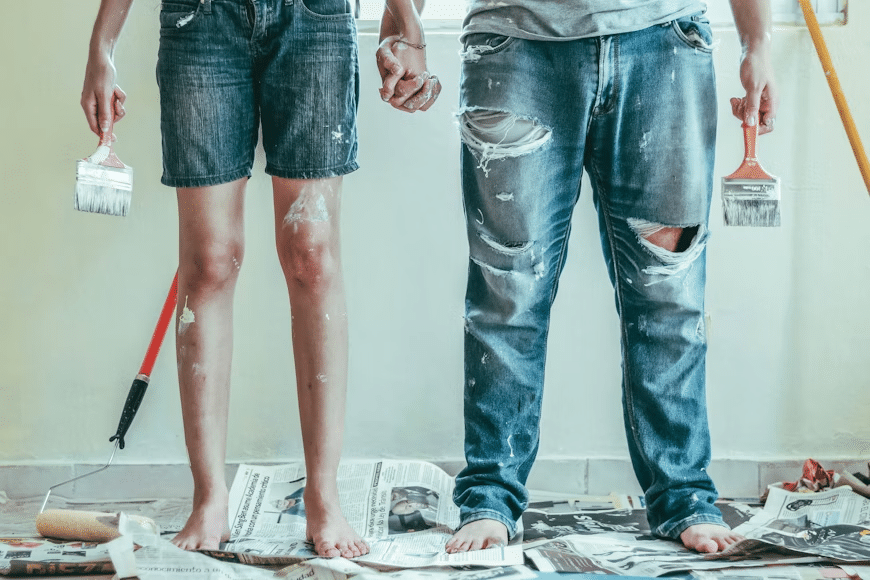 image Two people holding hands, covered in paint, standing barefoot on newspapers, ready for home DIY with painting tools. | Sky Rye Design Two people holding hands, covered in paint, standing barefoot on newspapers, ready for home DIY with painting tools.