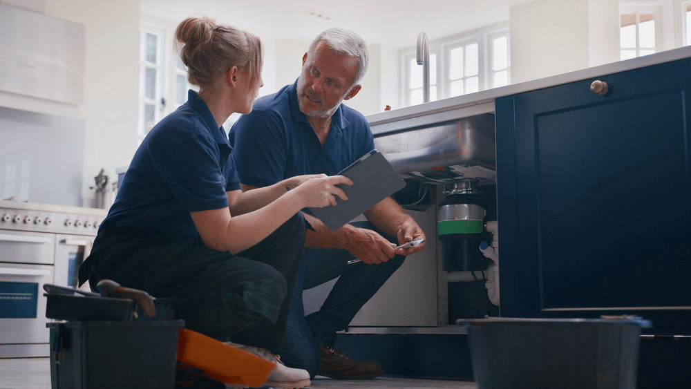 Two plumbers discussing kitchen sink repairs while checking under a cabinet.