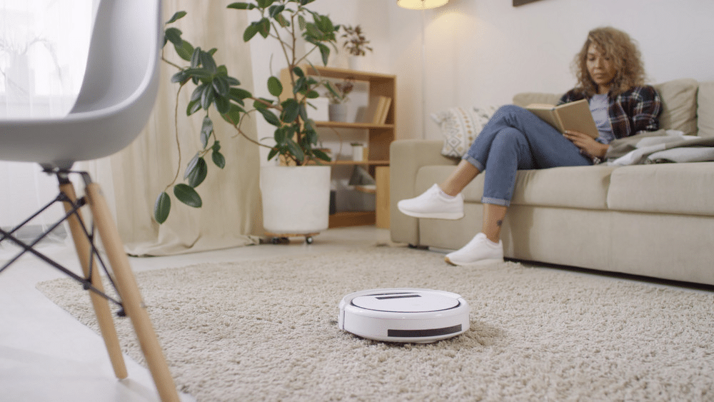 Woman reading on sofa while robot vacuum cleans carpet in modern living room.