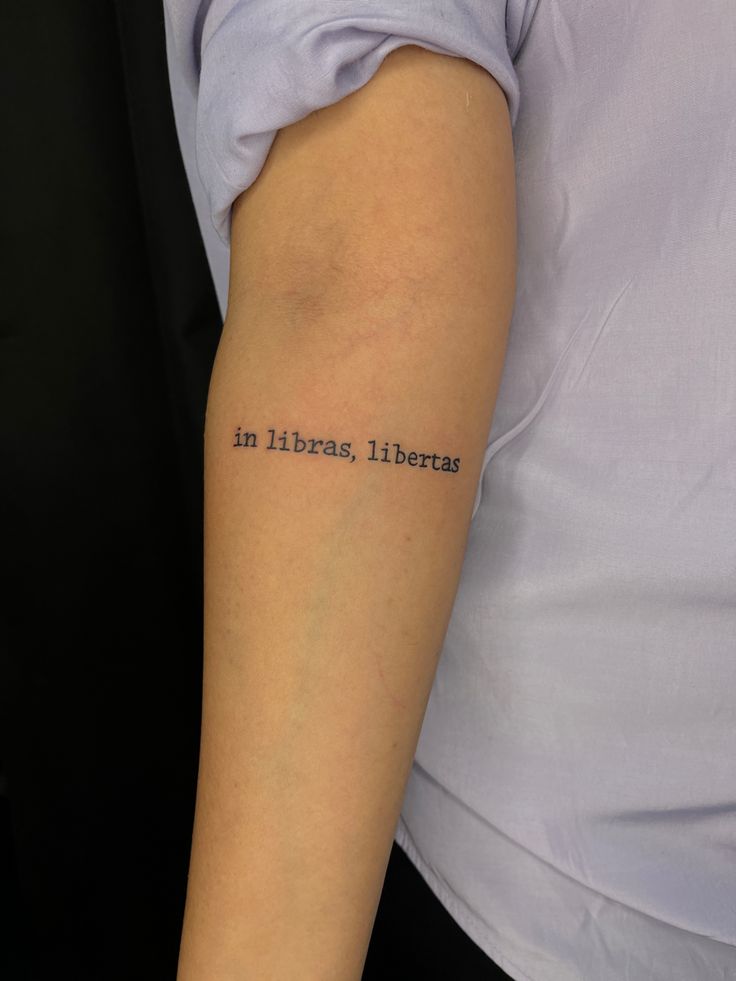 Close-up of an arm with the phrase in libras, libertas tattooed in simple black font, on light skin.