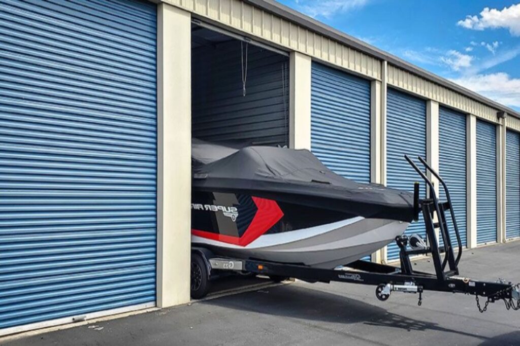 indoor-vs-outdoor-boat-storage-in-storage-unit Boat stored in a self-storage unit with blue shutters, partially outside on a trailer under a clear sky. | Sky Rye Design Boat stored in a self-storage unit with blue shutters, partially outside on a trailer under a clear sky.