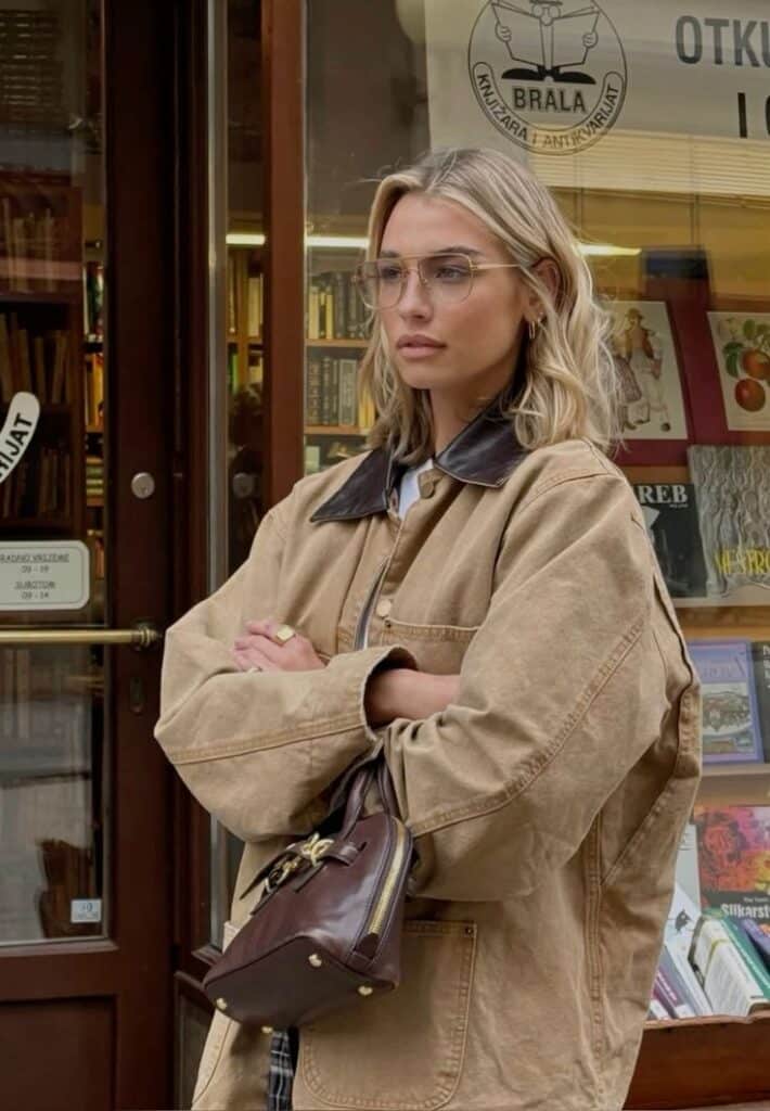 Trendy woman with glasses in a tan jacket stands outside a bookstore holding a brown bag.
