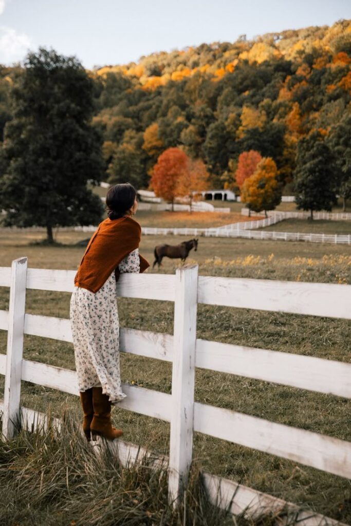Woman in a floral dress enjoys autumn scenery on a farm with horse and colorful trees.