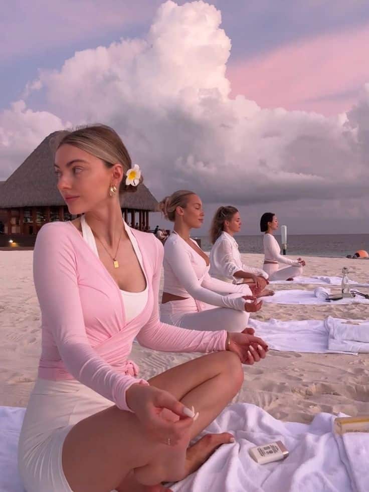 lizarudkevich Women meditating on a beach at sunset, wearing pastel clothing and seated on white towels, serene seascape in the background. | Sky Rye Design Women meditating on a beach at sunset, wearing pastel clothing and seated on white towels, serene seascape in the background.