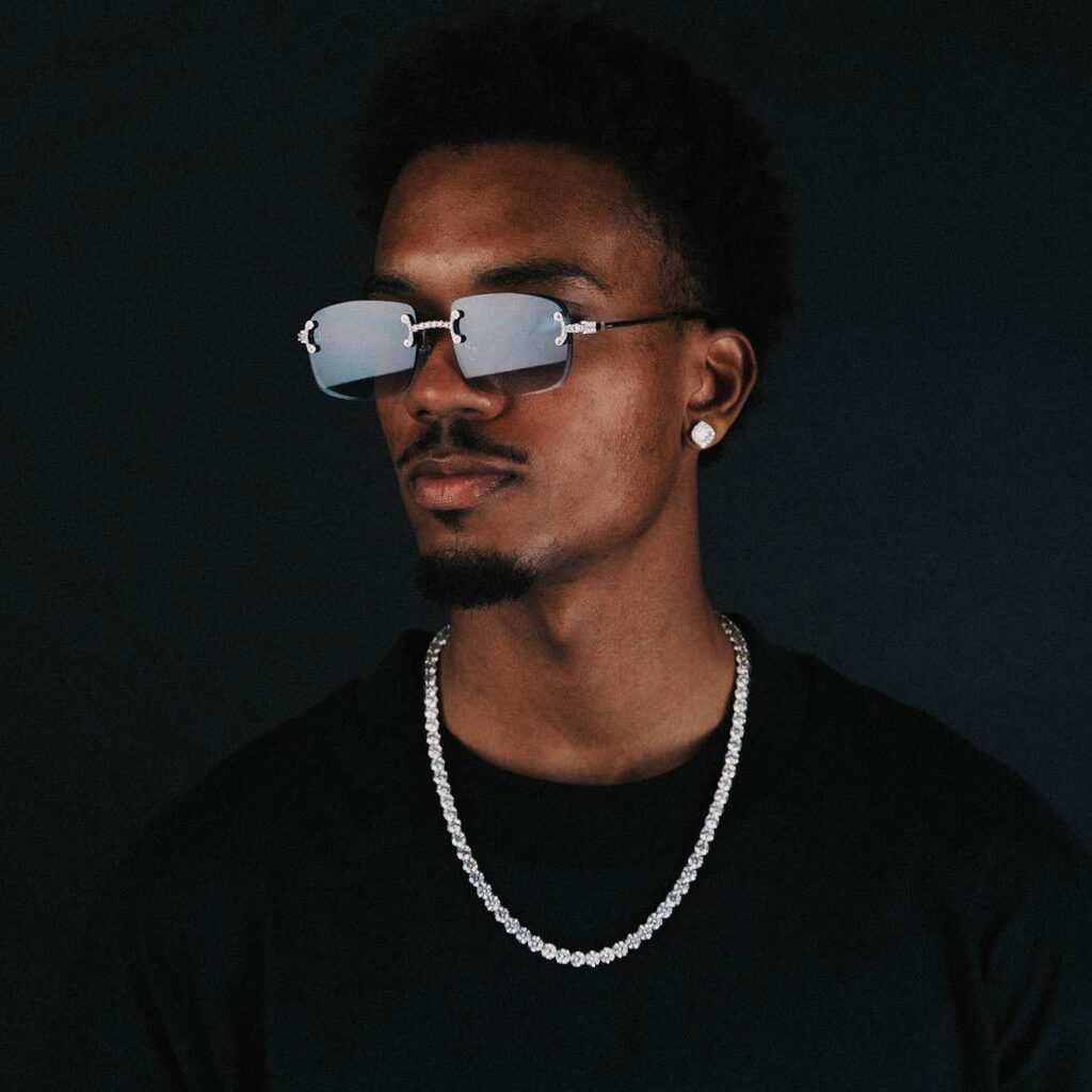 Man wearing stylish sunglasses and jewelry against a dark background.