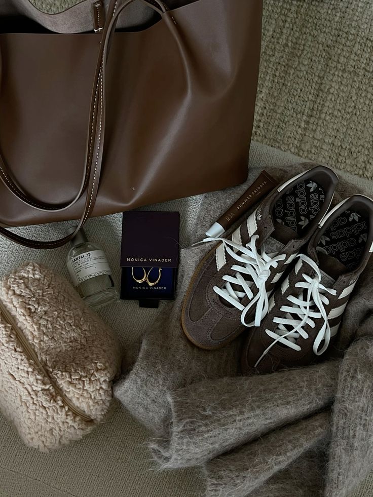 Brown leather bag with sneakers, jewelry box, perfume, cozy blanket, and a shearling pouch on a textured surface.
