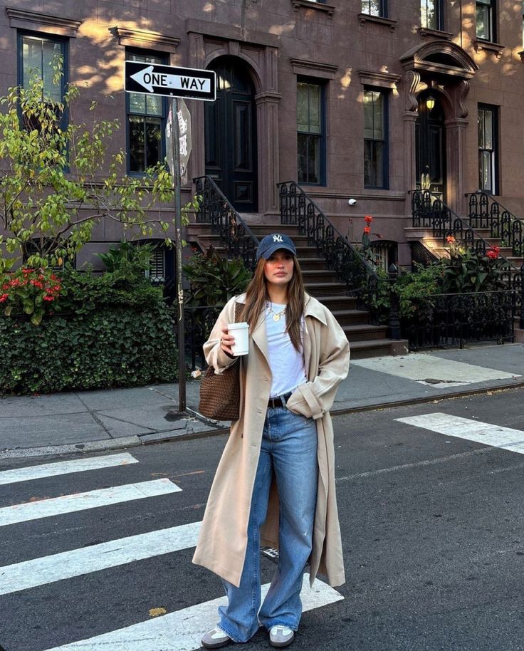 Woman in casual outfit with coffee on urban street, near brownstone building and one-way sign.