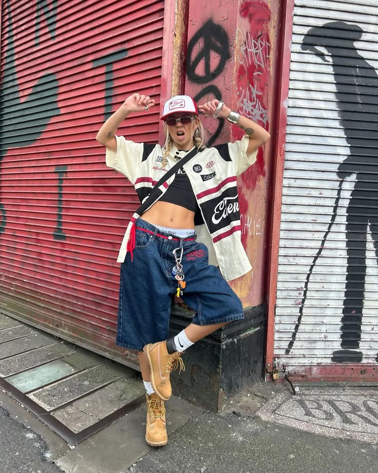 Person in streetwear poses playfully against a graffiti-covered wall, showcasing urban fashion with denim shorts and a cap.