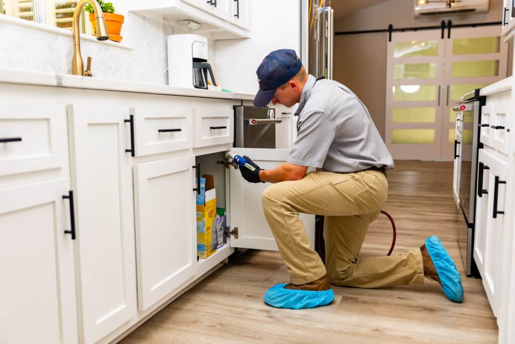 Maintenance worker fixing under-sink plumbing in a modern kitchen with white cabinets and wood flooring.