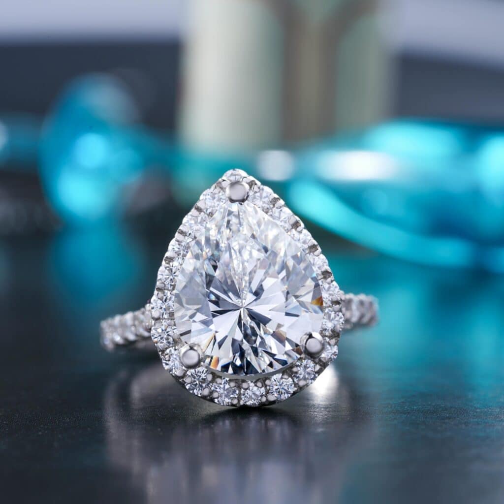 pexels-the-glorious-studio-3584518-6358515 Pear-shaped diamond ring with halo setting on reflective surface, elegant and sparkling with turquoise accents in background. | Sky Rye Design Pear-shaped diamond ring with halo setting on reflective surface, elegant and sparkling with turquoise accents in background.