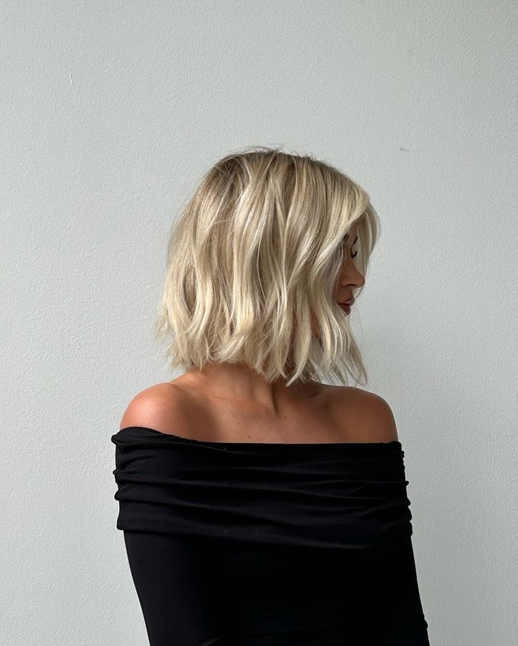 Blonde woman with wavy bob hairstyle wearing off-shoulder black top, looking to the side against a neutral background.
