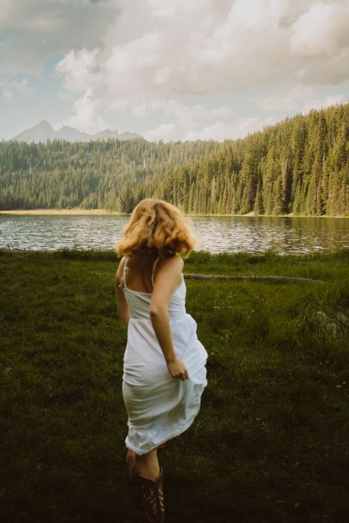 Woman in white dress running towards a scenic lake with forested hills and cloudy sky.