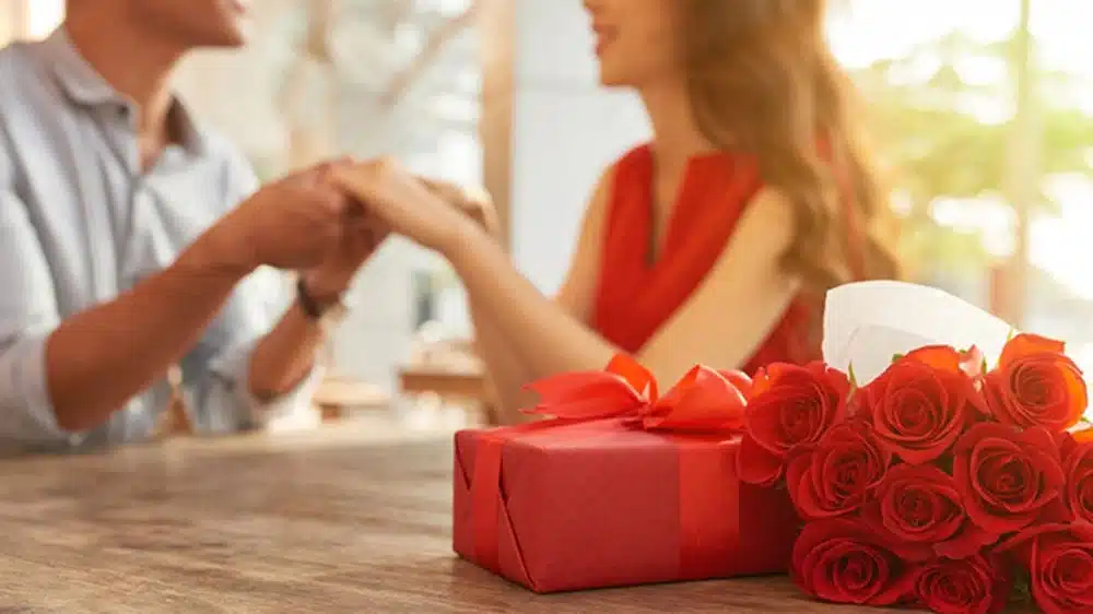 romantic-anniversary-gift-ideas_c92c0631-c0bf-41da-946b-af7d3af559e6 Couple holding hands in a romantic setting with red roses and a gift on the table. | Sky Rye Design Couple holding hands in a romantic setting with red roses and a gift on the table.
