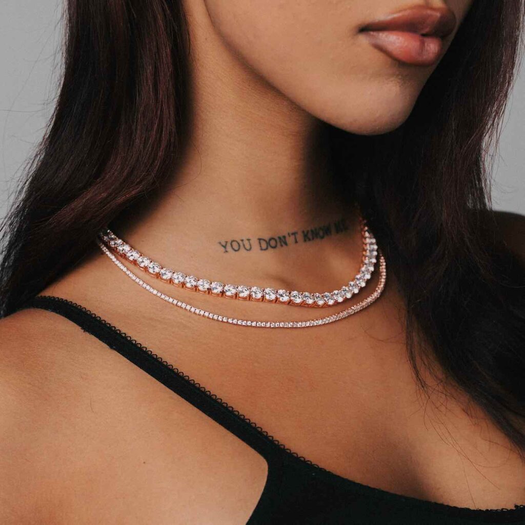 Close-up of a woman wearing layered diamond necklaces and a tattoo on her neck, against a neutral background.