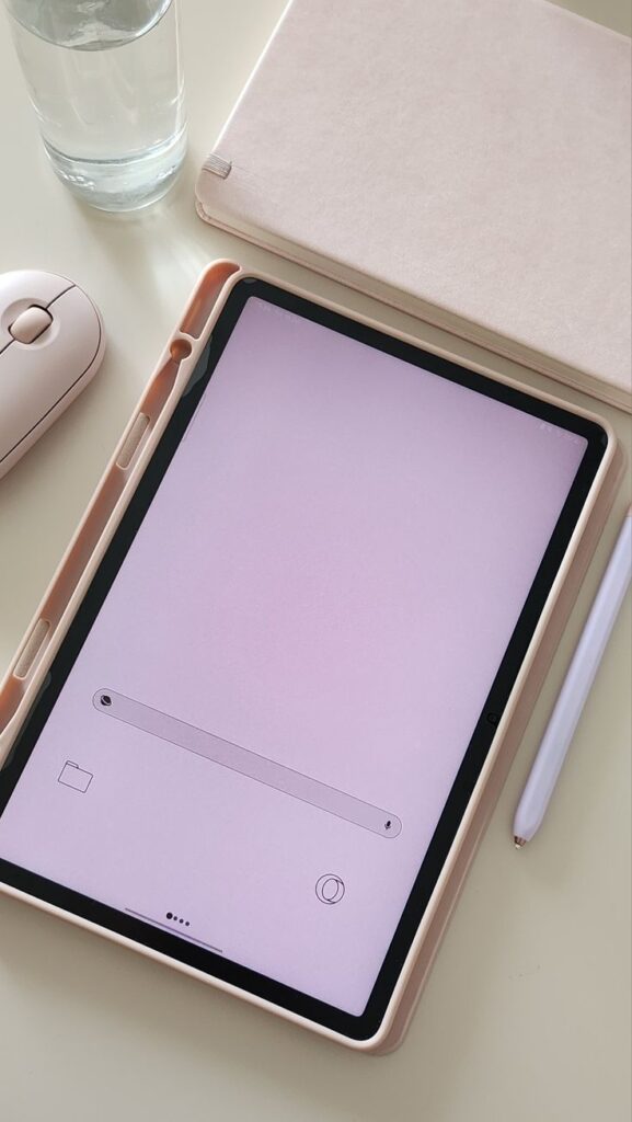 Tablet with pink screen, stylus, and mouse on desk next to notebook and glass of water. Minimalist workspace setup.