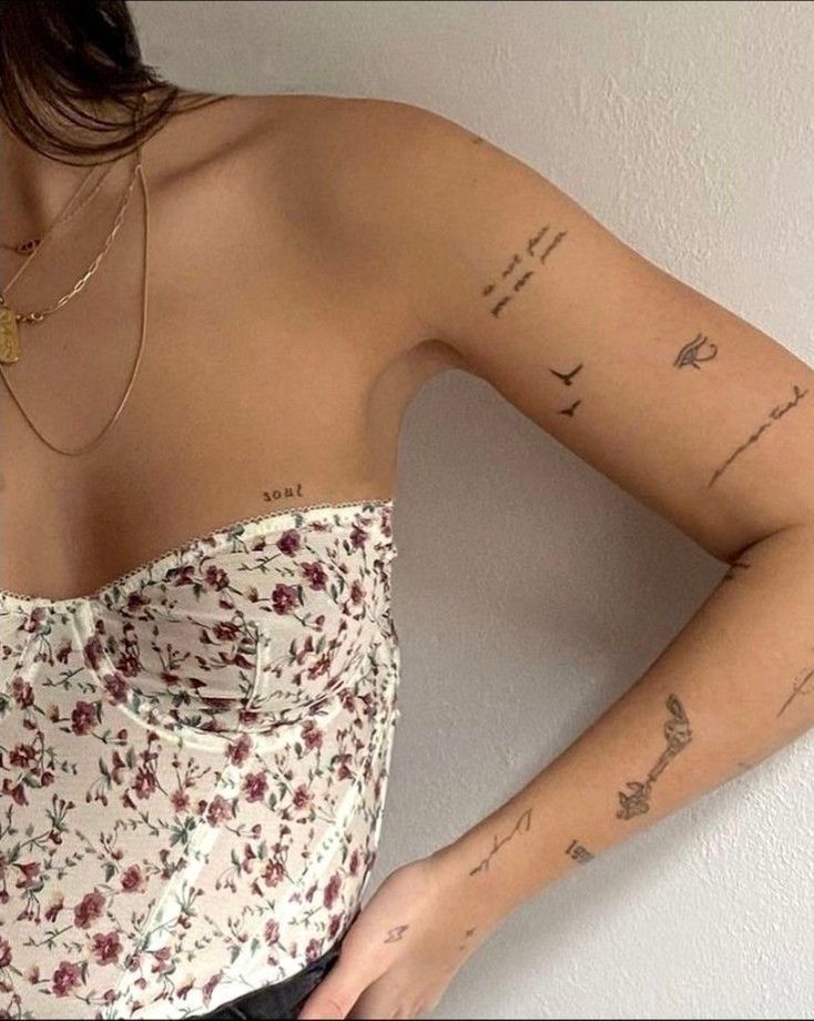 Woman in floral corset with delicate tattoos on arms and neckline, wearing layered gold necklaces.