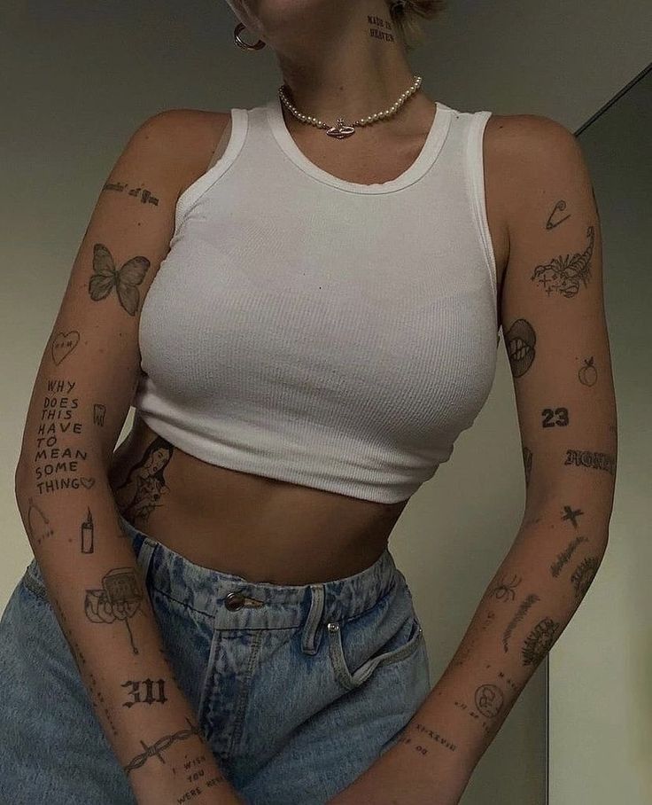 Person in a white tank top and jeans showcasing a variety of tattoos on arms and torso.