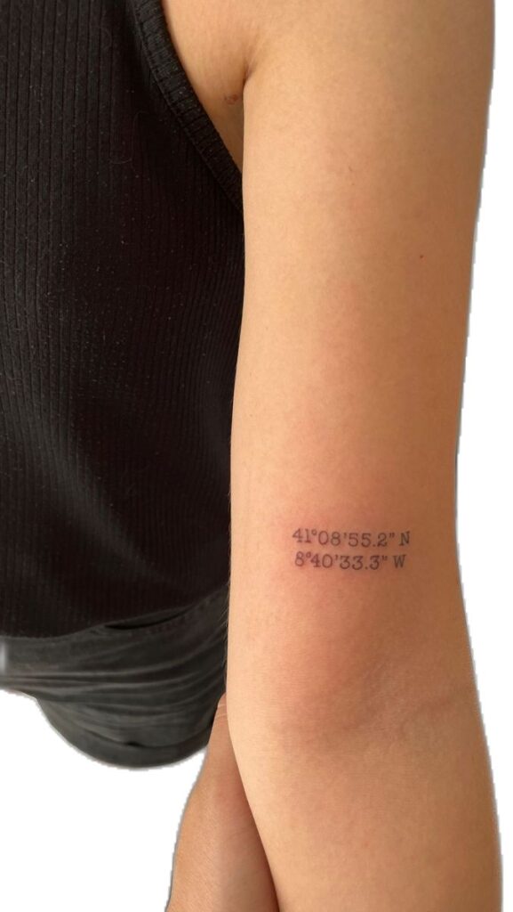 Tattoo of geographical coordinates on a person's arm, black tank top visible.