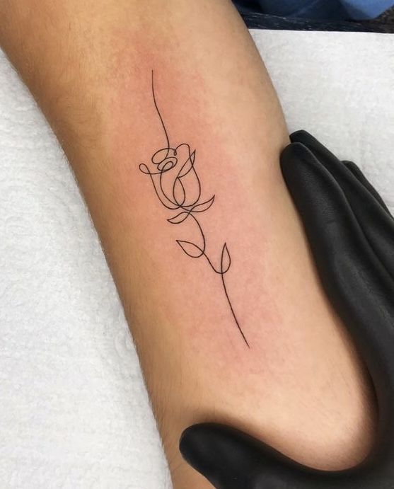 Minimalist rose line tattoo on forearm, held by gloved hand, on a light background.