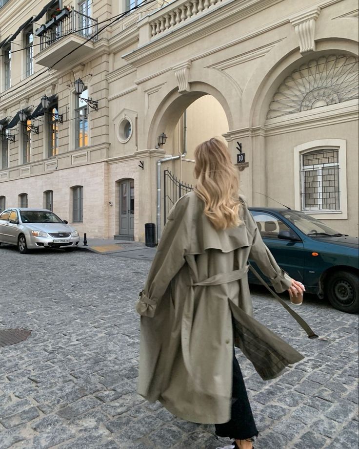 Woman in trench coat walking on cobblestone street near historic building.