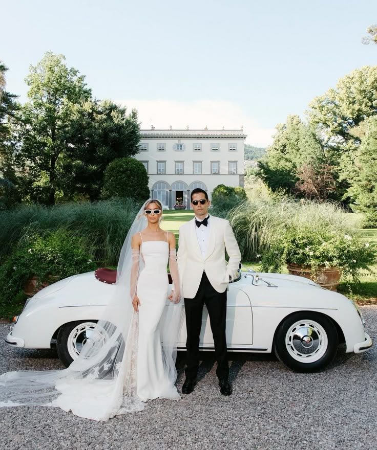 _ Bride and groom in elegant attire pose by vintage car in front of scenic estate garden. | Sky Rye Design Bride and groom in elegant attire pose by vintage car in front of scenic estate garden.