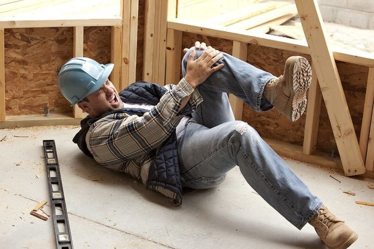 Construction worker in hard hat lying on floor, holding knee in pain. Workplace accident in unfinished building.