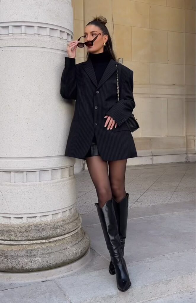 Woman in chic black blazer and boots, leaning on a column, wearing sunglasses and trendy accessories.