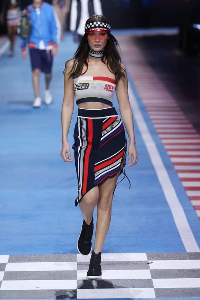 Fashion model on runway in sporty striped outfit and visor, blue and red theme, showcasing sleek modern style.