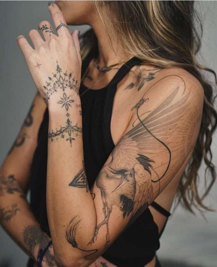 Woman with intricate tattoos on arm and hand, showcasing a crane design on arm, wearing a black sleeveless top.