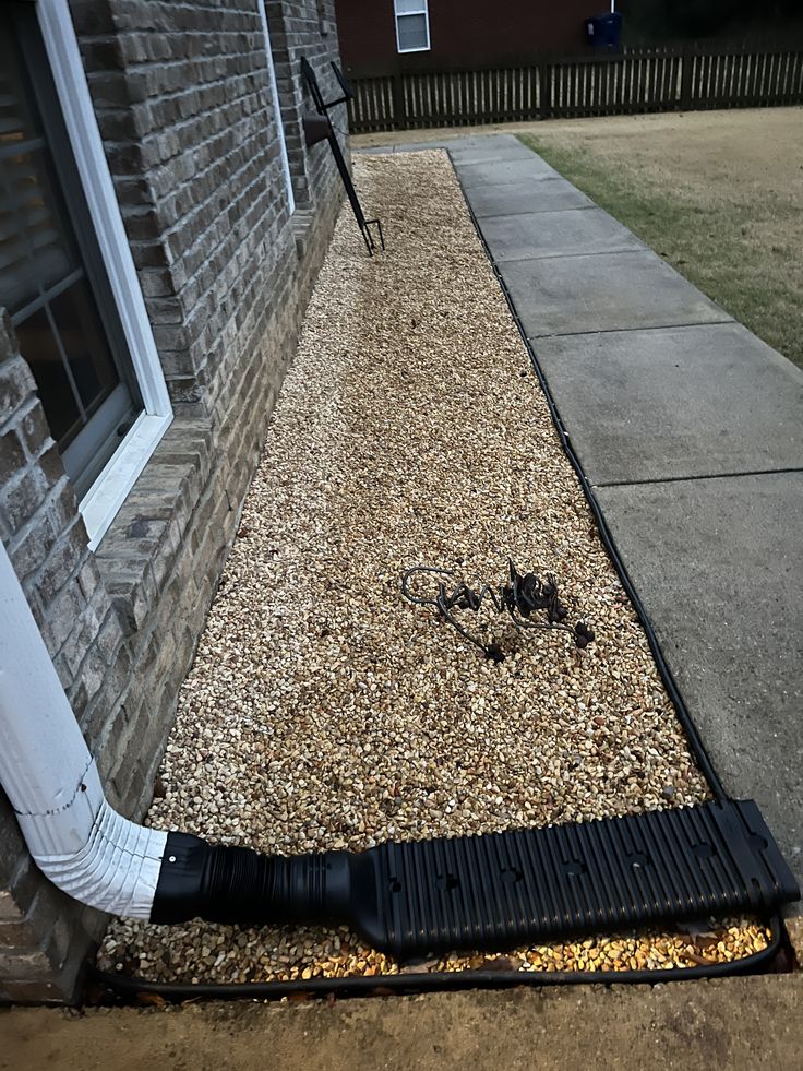 House exterior with brick wall, gravel landscaping, and drainage system beside a paved walkway.