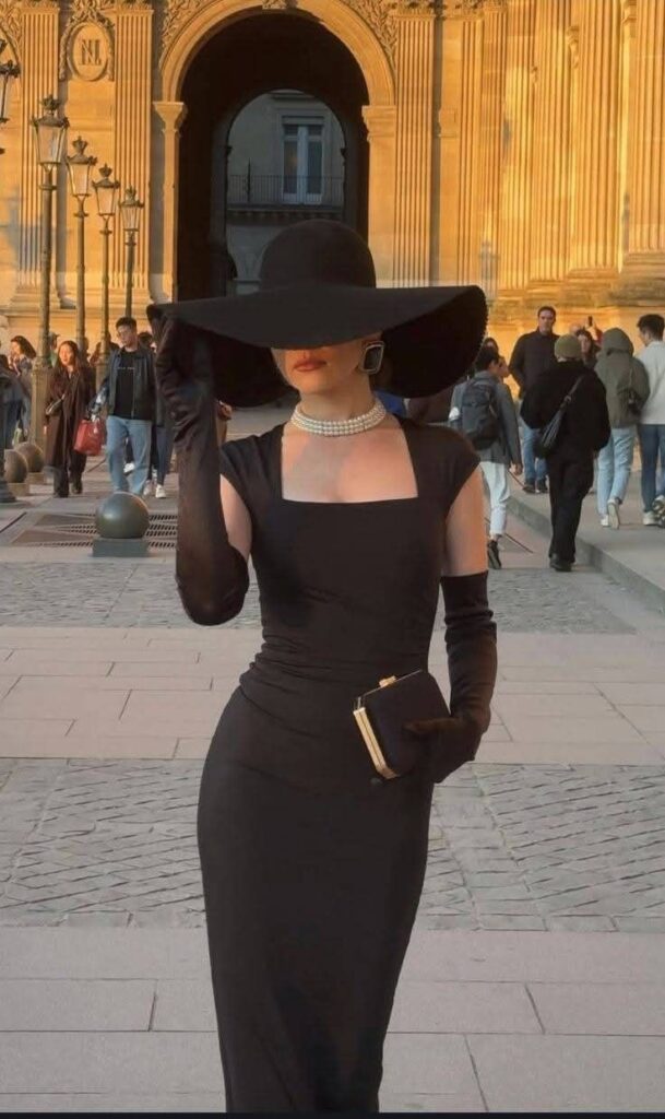 Stylish woman in elegant black dress and hat poses at historic archway, holding clutch in golden evening light.