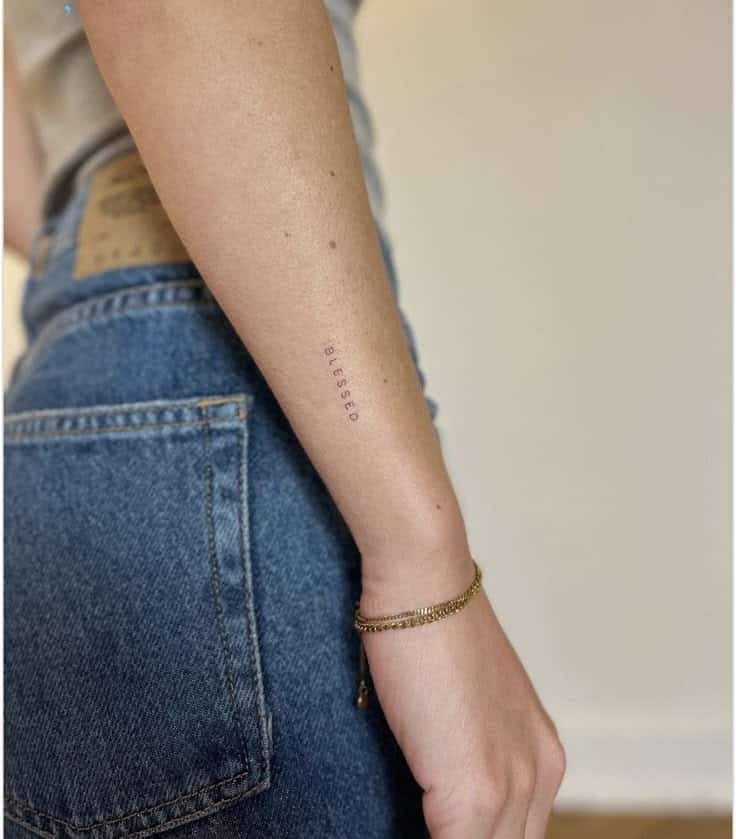Minimalist Blessed tattoo on forearm with gold bracelet, wearing denim jeans and beige top.