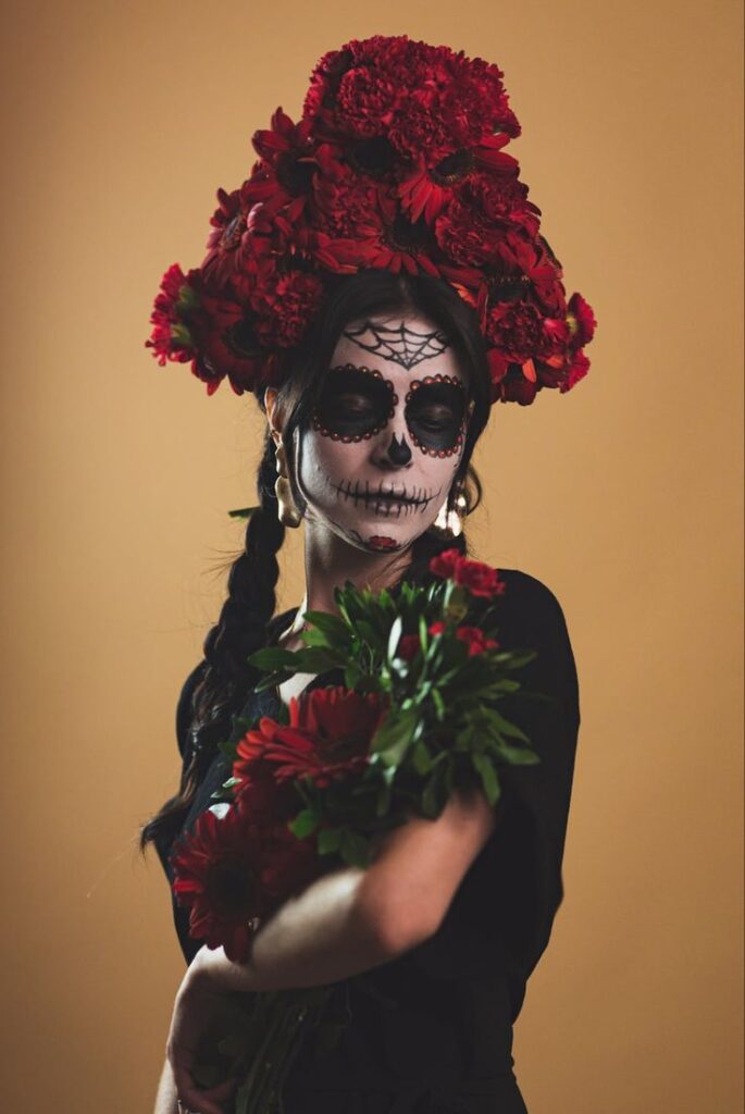 Woman in Día de los Muertos makeup, adorned with red flower headdress, holding bouquet, beige background.